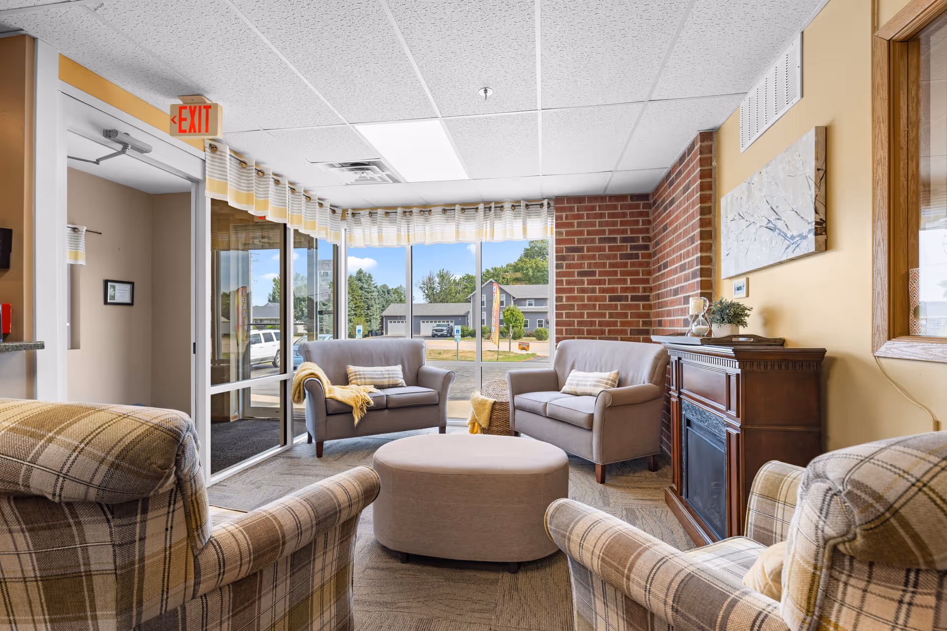 A cozy sitting area in an assisted living facility with two plaid armchairs, two gray loveseats, a round ottoman in the center, a wooden electric fireplace, and large windows showing an outdoor view. The walls are painted beige with a brick accent wall, and there is a painting above the fireplace. A glass door with an exit sign is visible on the left.
