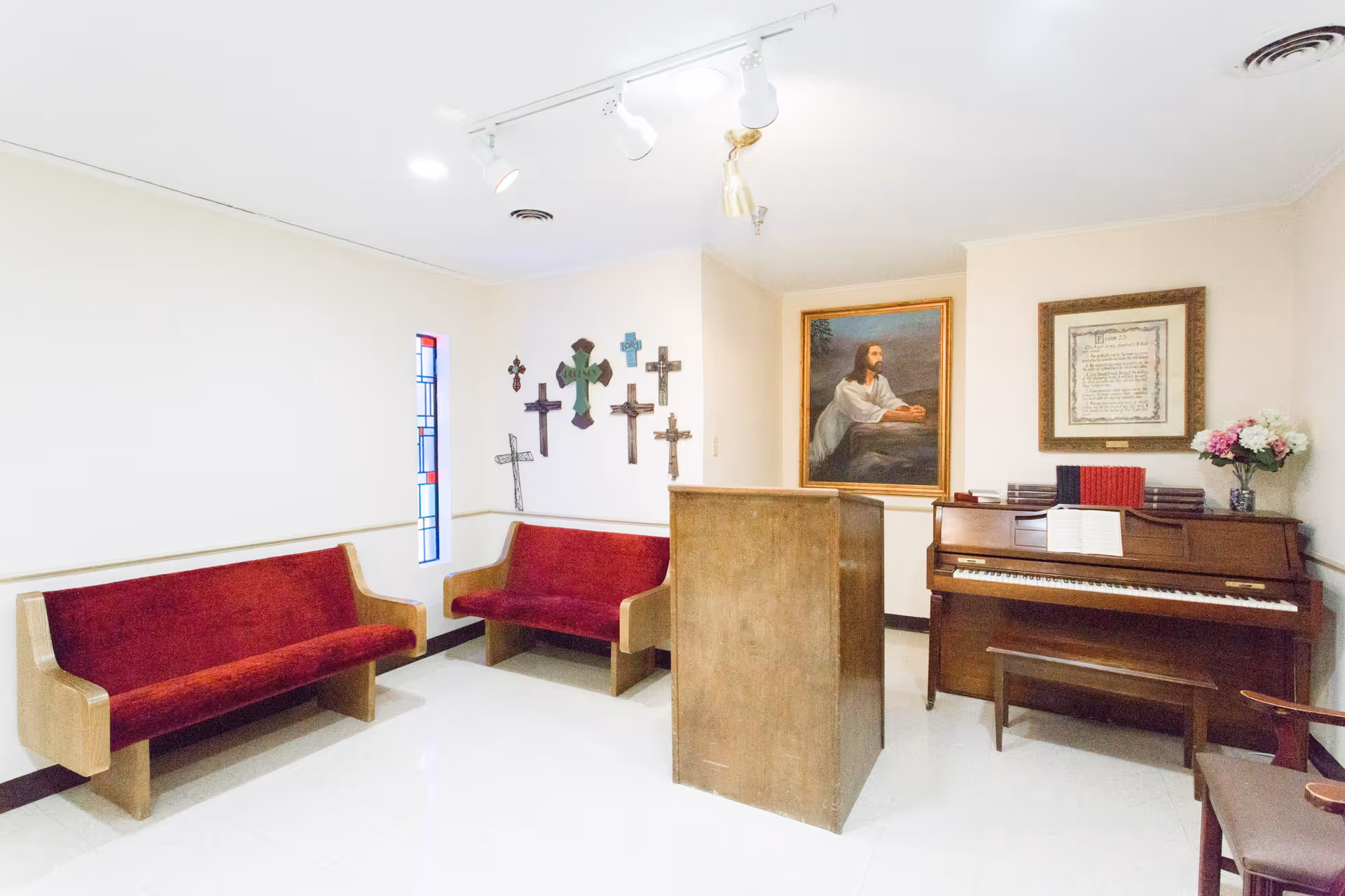 A small chapel room with two wooden pews with red cushions, a wooden podium, a piano with sheet music, and religious wall decorations including crosses and framed pictures.