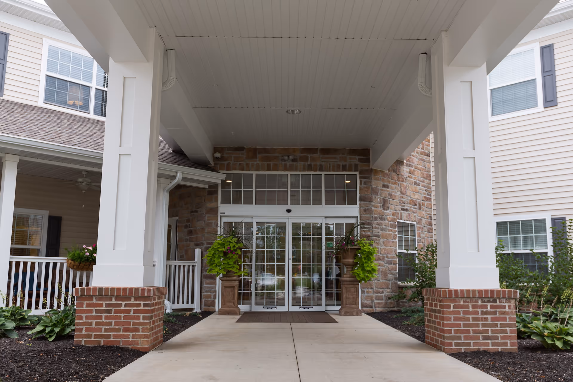 Entrance to a building with a covered walkway supported by white columns with brick bases. The entrance has glass double doors framed by stone walls and flanked by two large planters with green plants. The building exterior features beige siding and multiple windows with white trim and dark shutters.