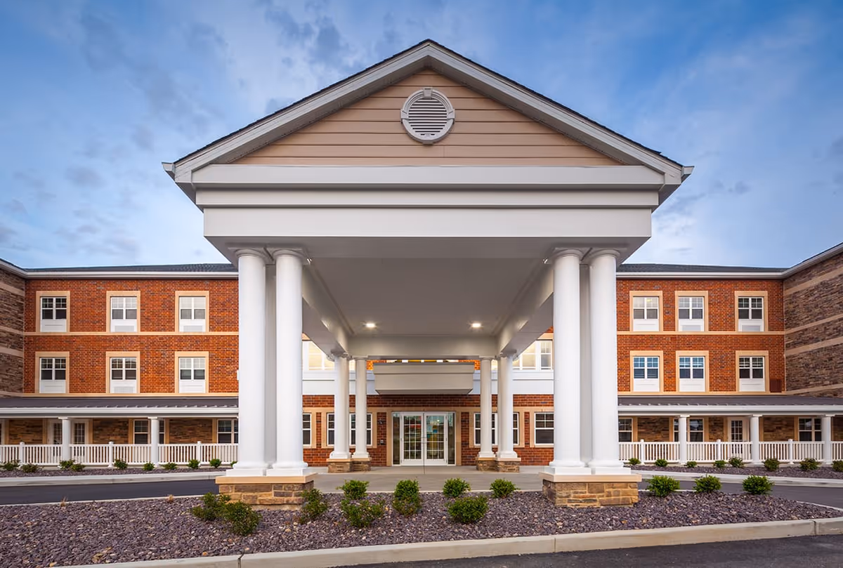 Front exterior view of Cedarhurst Senior Living of St. Charles building with a covered entrance supported by white columns, brick walls, multiple windows, and landscaped bushes under a partly cloudy sky.