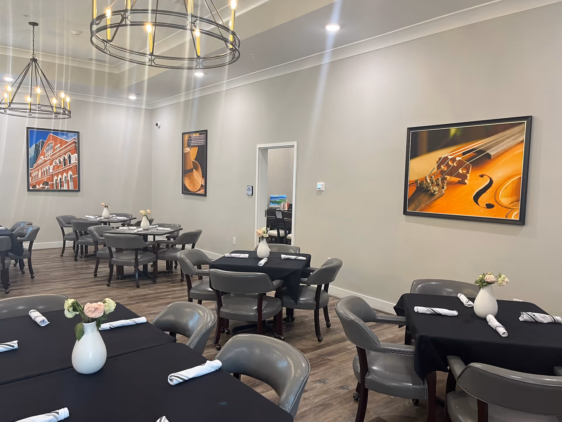 A dining room with several tables covered in black tablecloths, each set with rolled white napkins and small white vases holding flowers. The room has gray chairs around the tables, wooden flooring, and light gray walls adorned with framed pictures of musical instruments and a building. Two large circular chandeliers hang from the ceiling providing warm lighting.