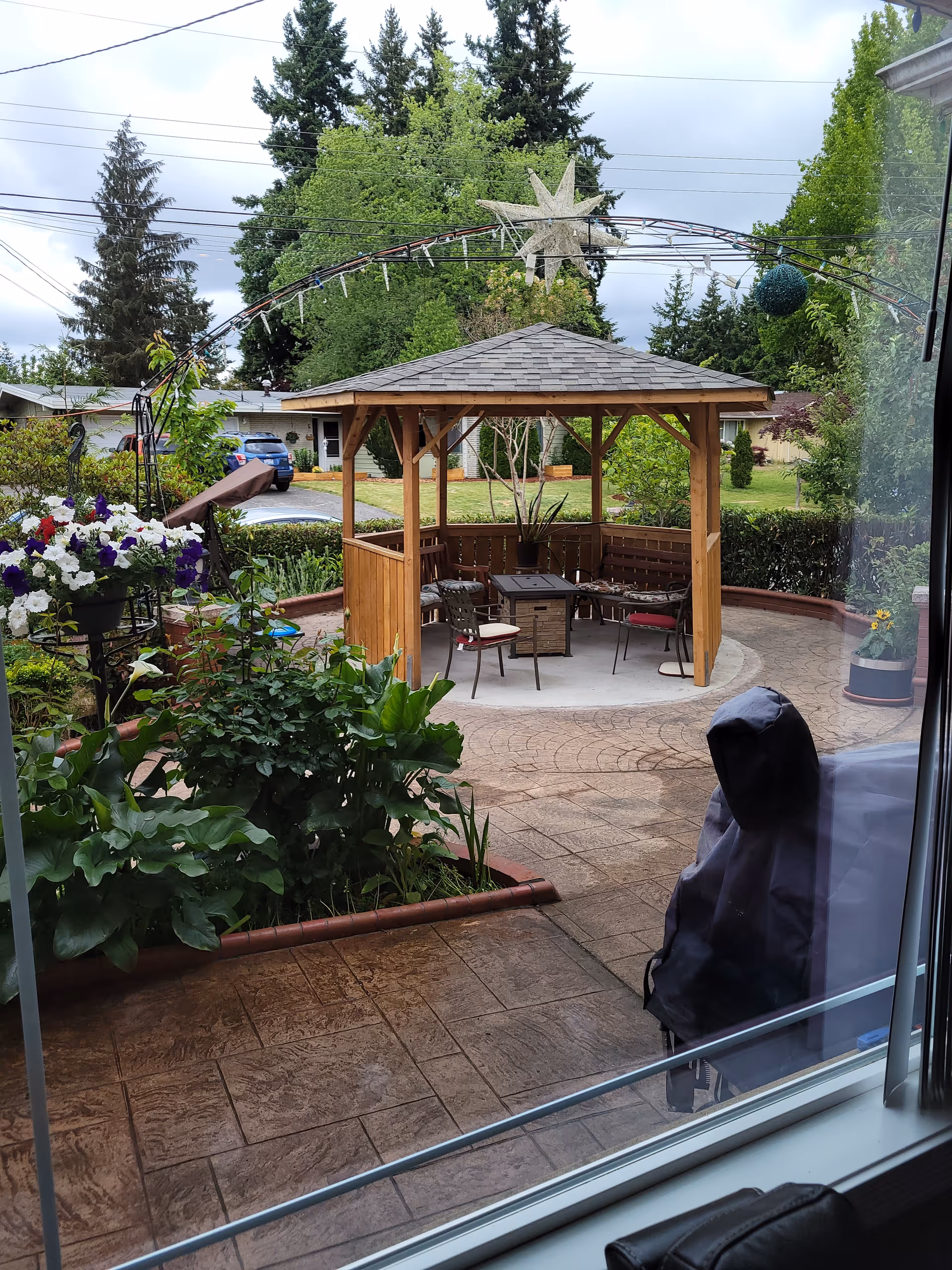 View through a window of an outdoor garden area featuring a wooden gazebo with a shingled roof, surrounded by chairs and a table. The garden has various green plants and flowers, a paved patio, and trees in the background under a cloudy sky.