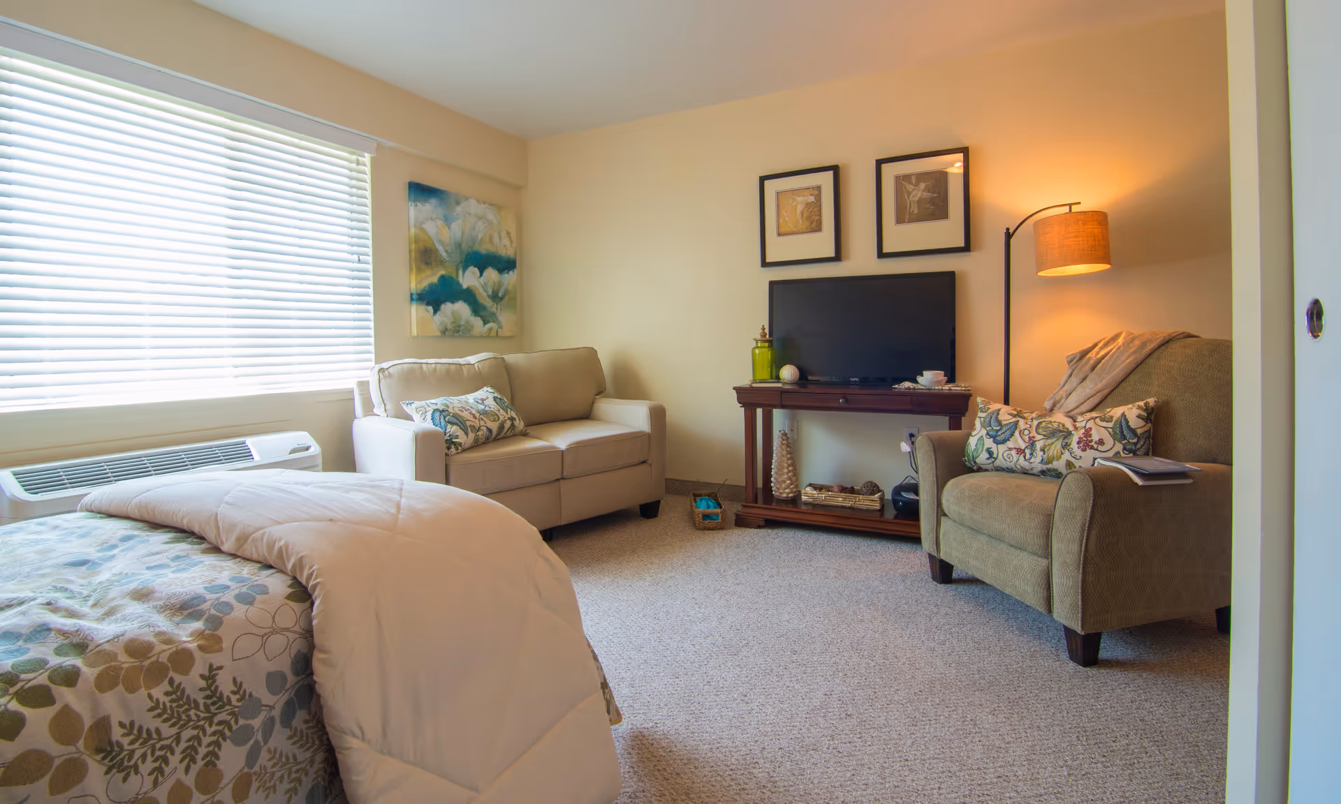 A cozy living area with a beige loveseat, a green armchair with a floral pillow, a wooden TV stand holding a flat-screen TV and decorative items, a floor lamp with a warm light, and a bed with a floral comforter partially visible in the foreground. A large window with white blinds lets in natural light, and two framed artworks hang above the TV.