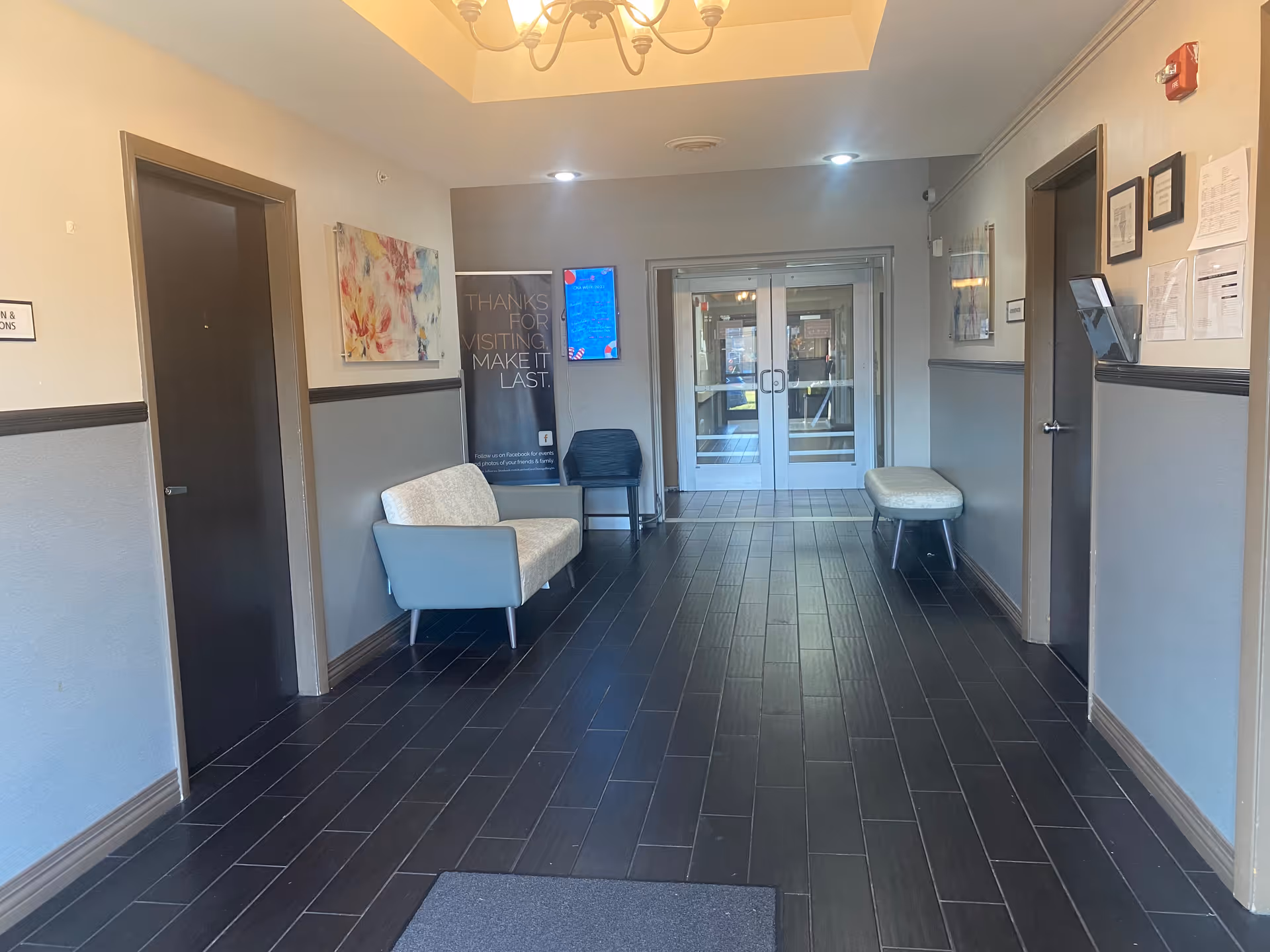 Interior hallway of a senior living facility with dark tiled floor, light gray walls with dark trim, two closed doors on each side, a white cushioned bench and a black chair on the left, a light cushioned bench on the right, a colorful abstract painting on the left wall, a banner with text 'Thanks for visiting, make it last' near the back, and double glass doors at the end of the hallway.