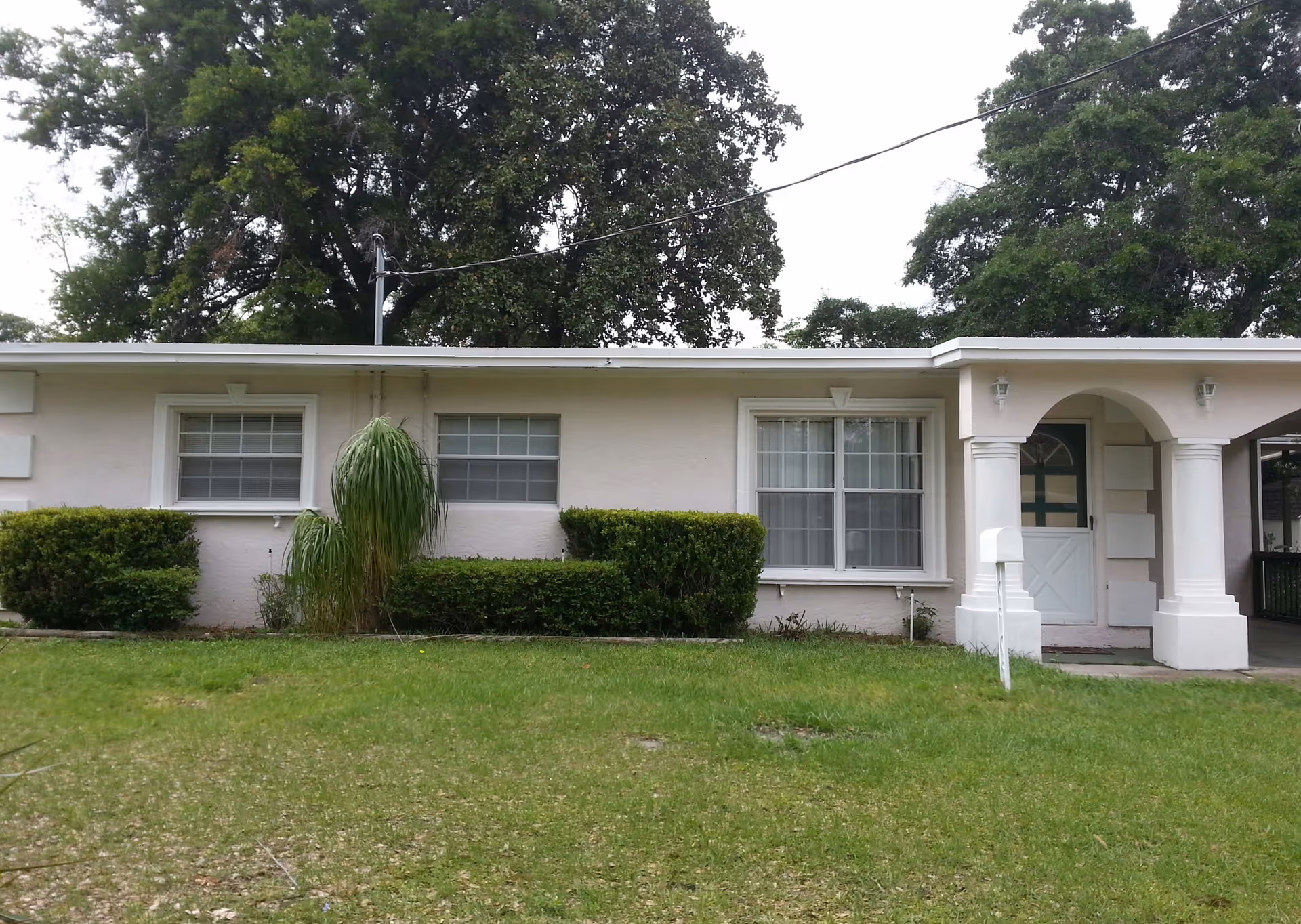 Single-story residential building with light-colored exterior walls, two large windows with white frames, a front door under an archway supported by two columns, surrounded by green bushes and a grassy lawn with large trees in the background.