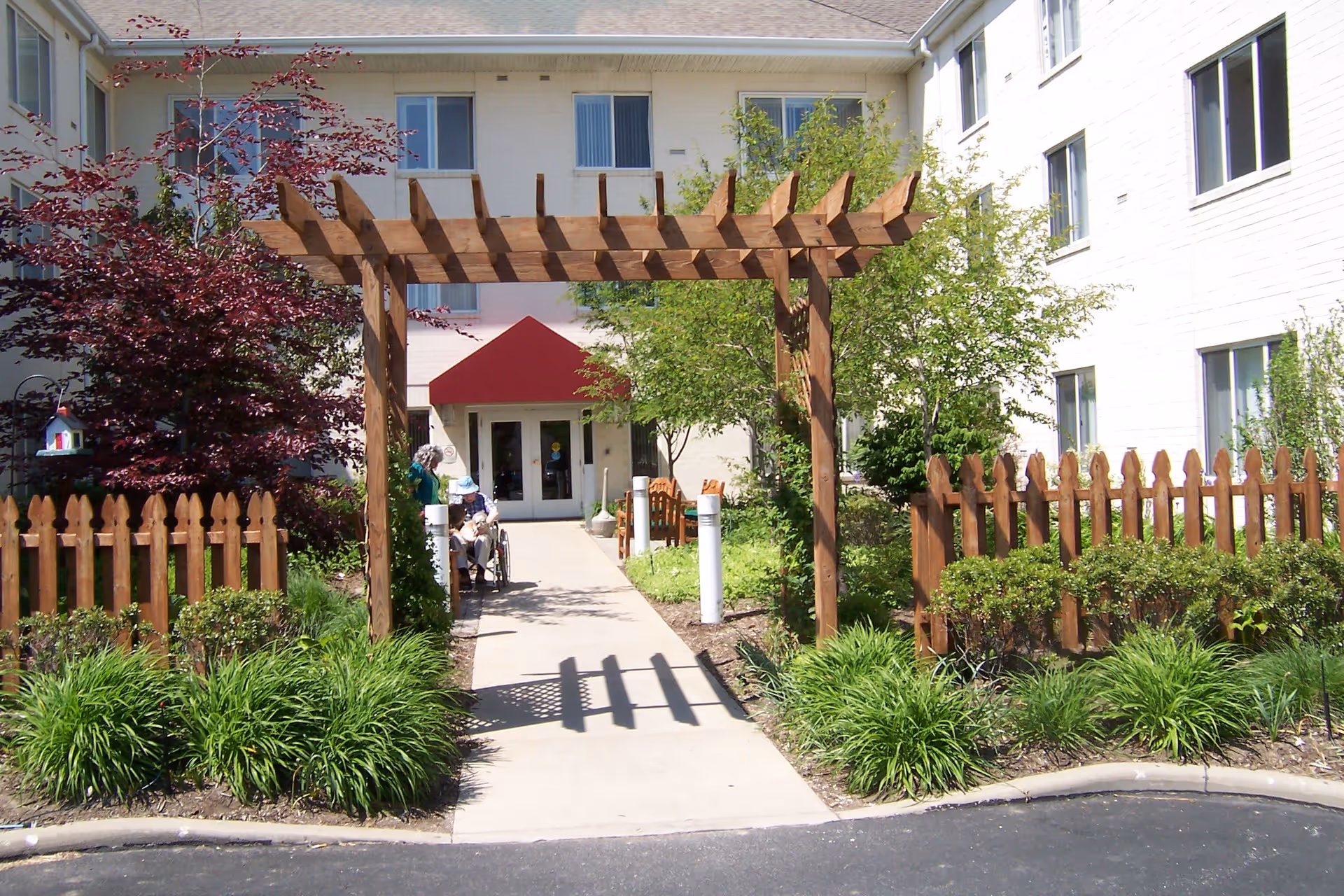 Entrance to a senior living facility with a wooden pergola over a concrete walkway, surrounded by green shrubs and trees. A wooden picket fence lines both sides of the walkway. Two elderly people, one in a wheelchair, are near the entrance which has a red awning above the doors. The building is white with multiple windows.