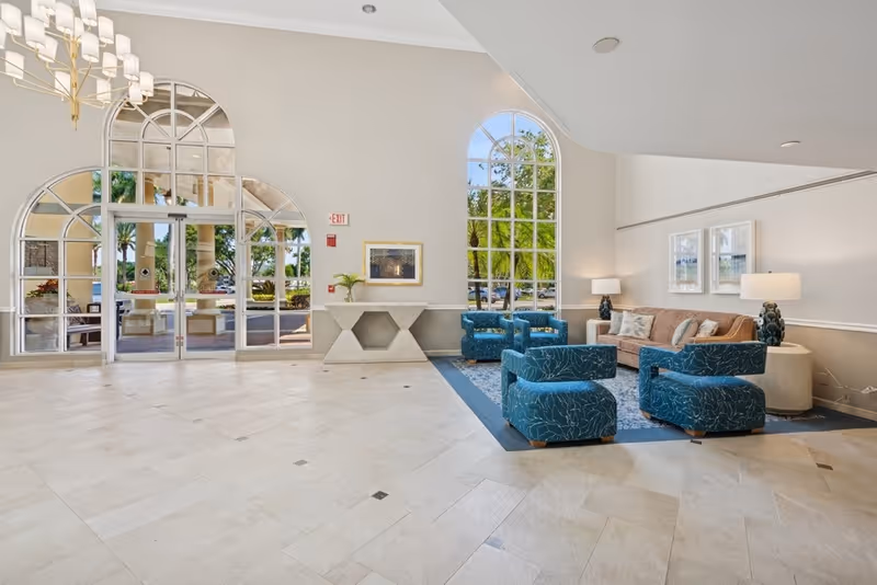Bright senior living lobby with blue armchairs and a sofa arranged on a rug beside large arched windows and glass entry doors.