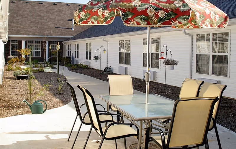 Outdoor patio area at Meadow Lakes with a glass table and six beige chairs under a colorful floral umbrella. The patio is adjacent to a white building with multiple windows and a curved concrete walkway surrounded by garden beds with some plants and a green watering can.