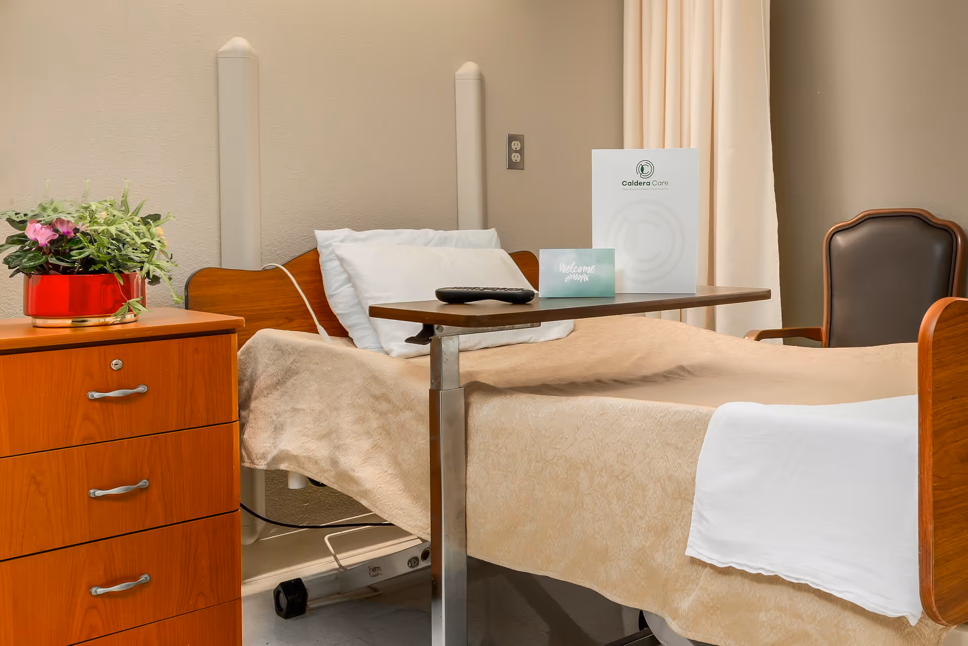 A hospital-style bed with beige bedding and white pillows in a room. There is a wooden bedside table with a red pot containing green and purple plants. A brown chair is positioned next to the bed. A small overbed table holds a remote control, a welcome card, and a Caldera Care brochure. The walls are beige, and there is a curtain in the background.