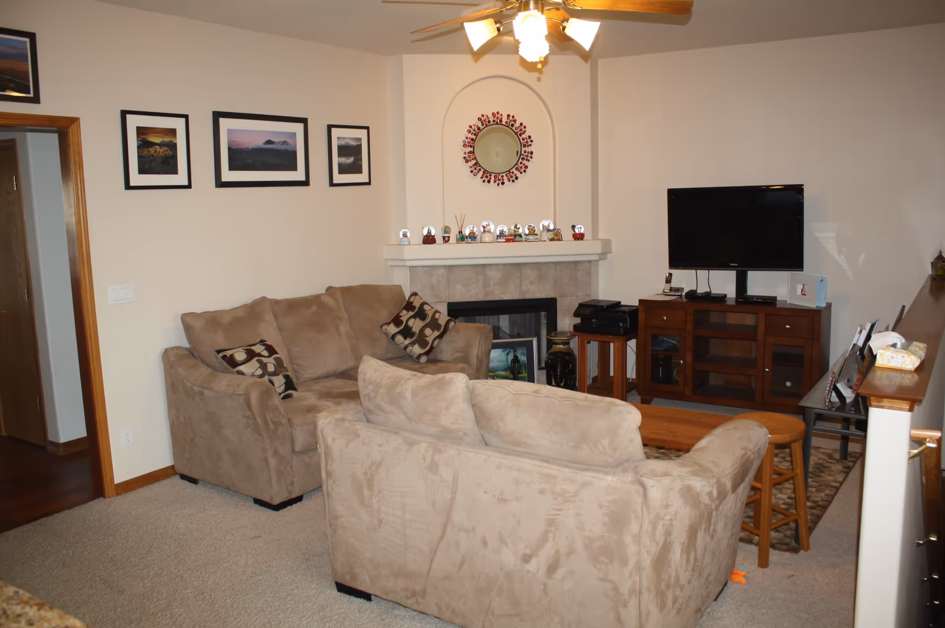 A cozy living room with two beige sofas facing a wooden TV stand with a flat-screen television. The room features a fireplace with decorative items on the mantle, a round mirror above it, and framed pictures on the wall. There is a ceiling fan with lights and a small wooden stool in front of the sofas.