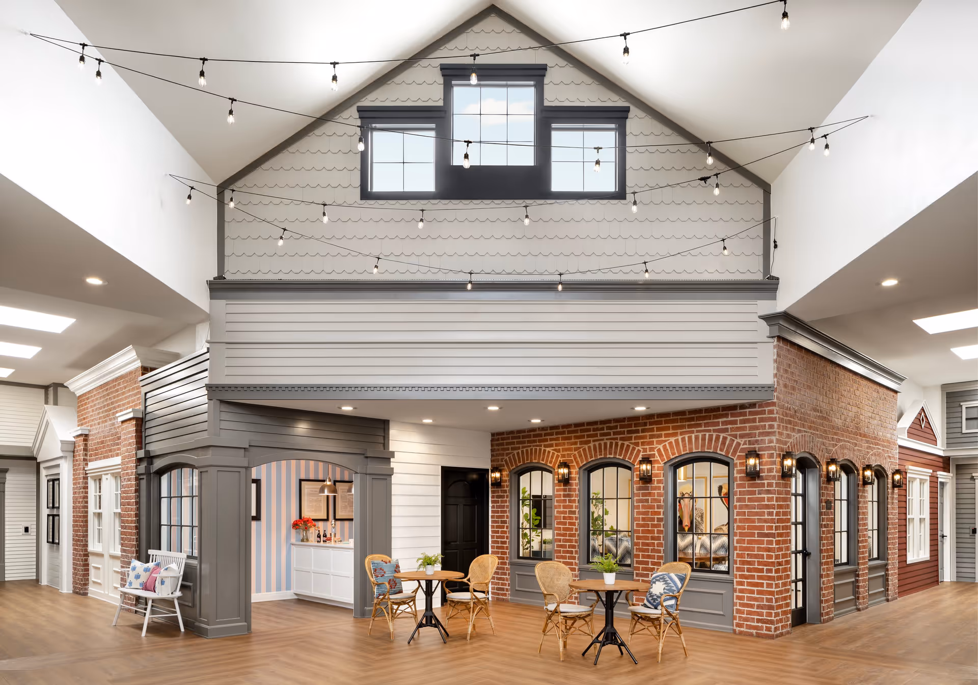 Interior common area styled like an indoor village with brick and clapboard storefront facades, string lights overhead, and small café tables and chairs.