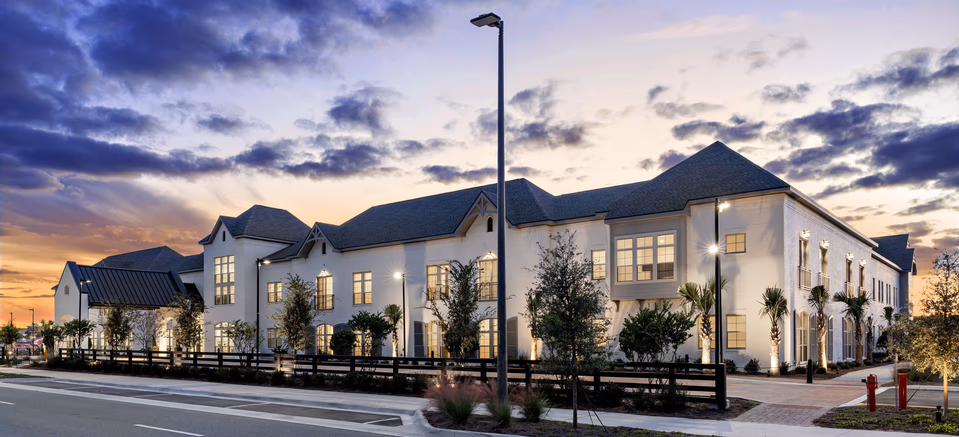 Exterior view of a large, two-story senior living facility building at dusk with lights illuminating the windows and landscaping. The building features a pitched roof, multiple windows, and is surrounded by trees and a wooden fence. The sky shows a colorful sunset with clouds.