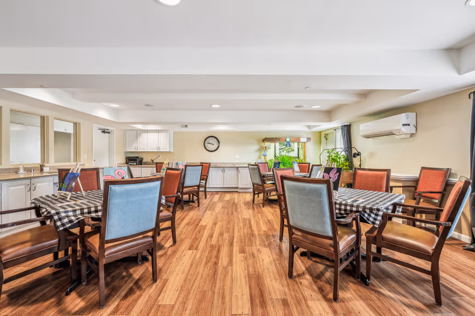 A bright and spacious dining room with several tables covered in black and white checkered tablecloths. Each table is surrounded by wooden chairs with cushioned seats and backs in various colors. The room has wood flooring, light-colored walls, a clock on the far wall, and a window with plants on the windowsill. There is also a white air conditioning unit mounted on the right wall and cabinets along the back wall.