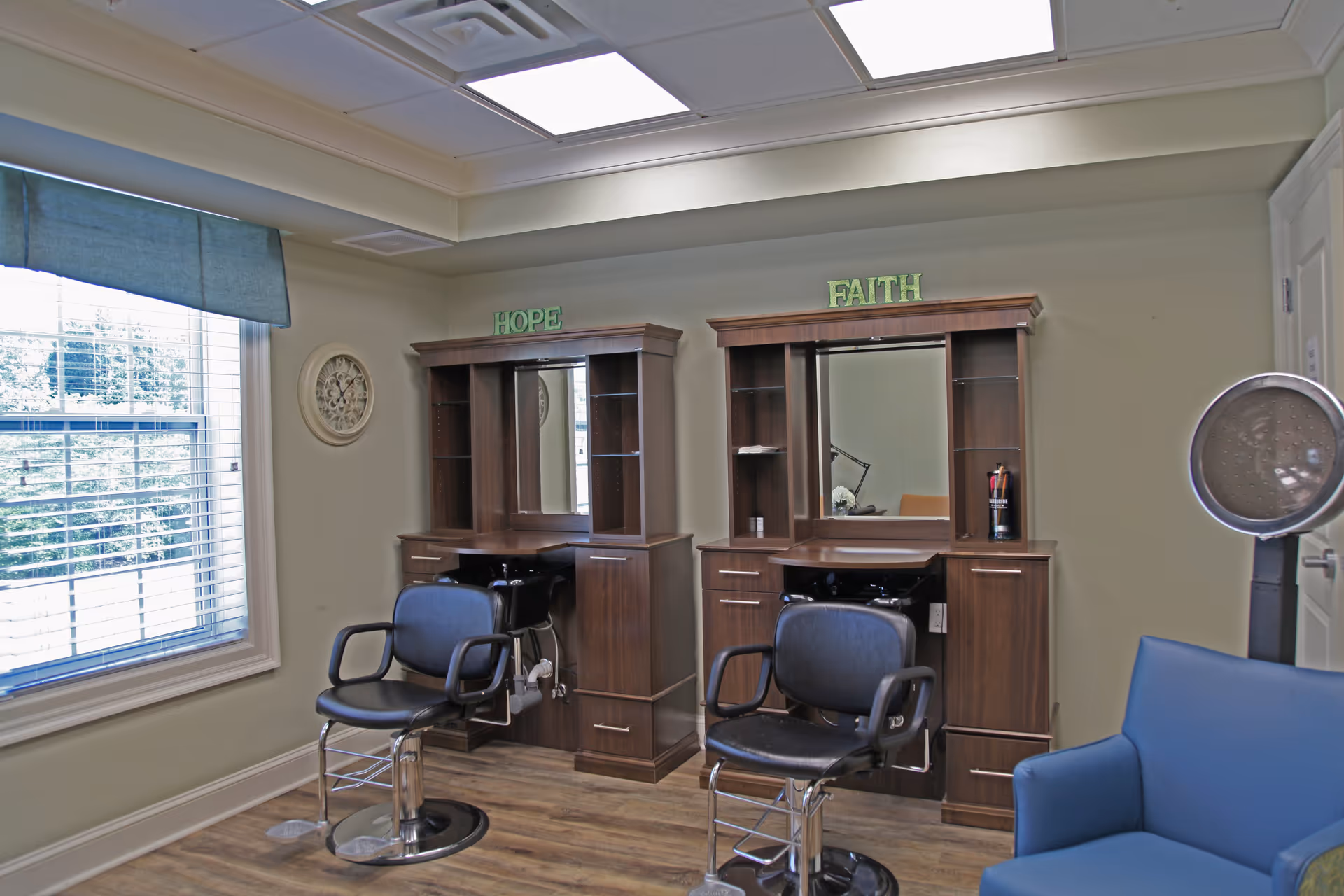 Interior view of a salon area with two styling stations labeled 'HOPE' and 'FAITH'. Each station has a mirror, shelves, and a black salon chair. There is a large window with blinds and a blue valance on the left side, and a blue armchair with a hair dryer on the right side. The room has light-colored walls and wood flooring.