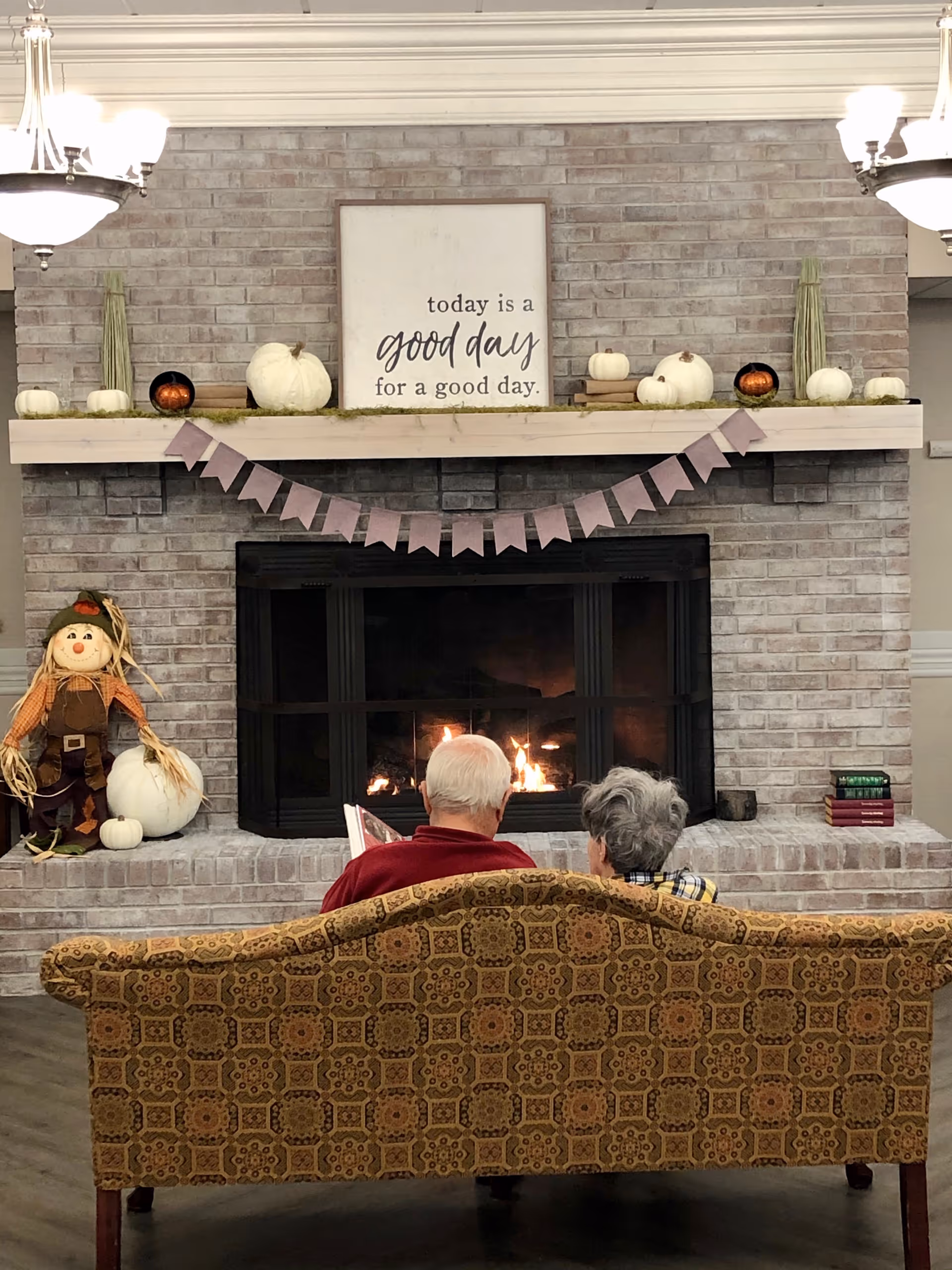 Two elderly people sitting on a patterned couch facing a lit fireplace in a cozy room. The fireplace mantel is decorated with white and orange pumpkins, books, and tall green plants. Above the mantel is a sign that reads 'today is a good day for a good day.' A small scarecrow decoration is placed to the left of the fireplace.
