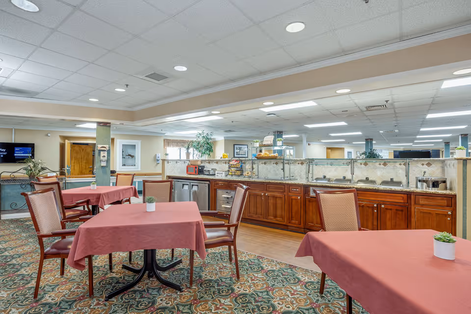 Interior view of a senior living facility dining area with tables covered in red tablecloths and chairs around them. The room features a patterned carpet, a long counter with wooden cabinets, and various kitchen appliances and food items on the counter. The ceiling has recessed lighting and there are plants and framed pictures on the walls.