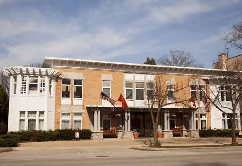 Two-story brick senior living facility with white accents, flags, and a front entrance.