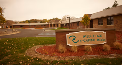 Exterior view of a single-story brick senior living facility with a landscaped sign reading "Medilodge of Capital Area" in the foreground.