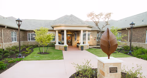 Front entrance of a brick memory care community with a covered entry, landscaped lawns, and a metal leaf sculpture on a pedestal.