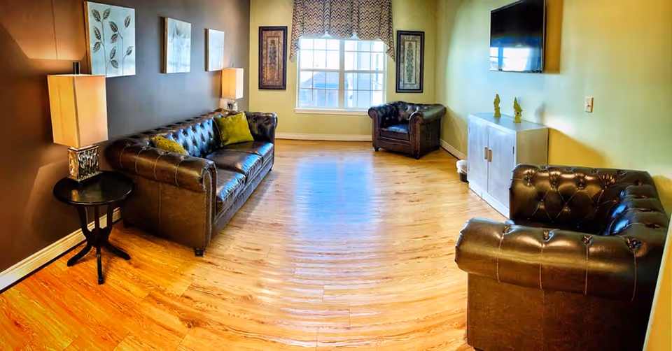 A cozy living room with wooden flooring, featuring a dark brown leather sofa with green cushions on the left, two matching leather armchairs on the right and near the window, a small round side table with a lamp, a white cabinet with two small decorative items on top, a wall-mounted flat screen TV, and a large window with patterned curtains letting in natural light.