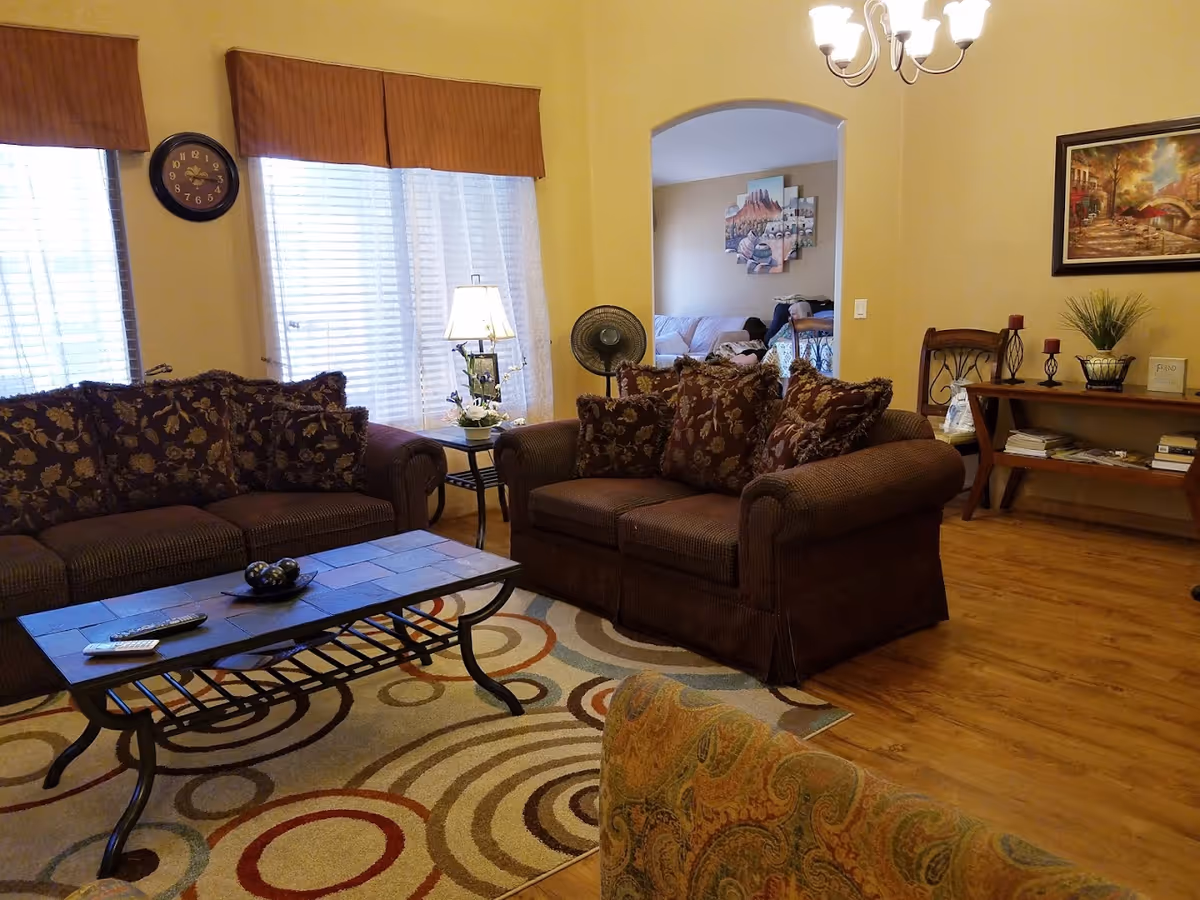 A cozy living room with two brown patterned sofas, a coffee table with remote controls and decorative balls, a colorful circular patterned rug, a side table with a lamp and flowers, a wall clock, and a console table with candles, books, and a plant. The room has wooden flooring, yellow walls, and a chandelier. An archway leads to another room with a couch and wall art.