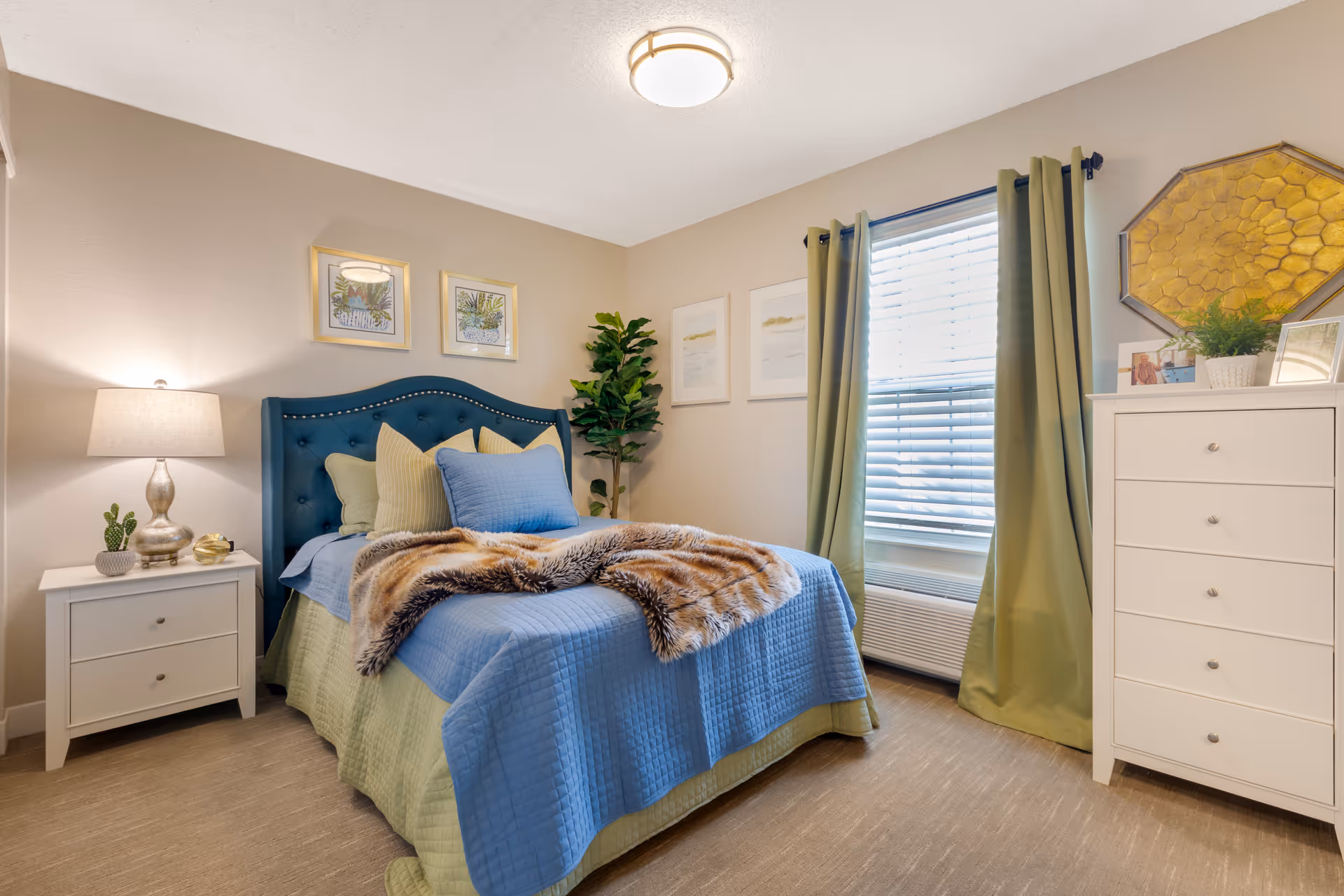 A cozy bedroom with a blue upholstered headboard bed dressed in blue and green bedding with a faux fur throw. There is a white nightstand with a lamp and small decorative items on the left side of the bed. On the right side, there is a white dresser with framed photos, a plant, and a decorative golden hexagonal wall piece above it. A window with green curtains and white blinds is next to the dresser. The walls are beige with framed artwork hanging above the bed and near the window.