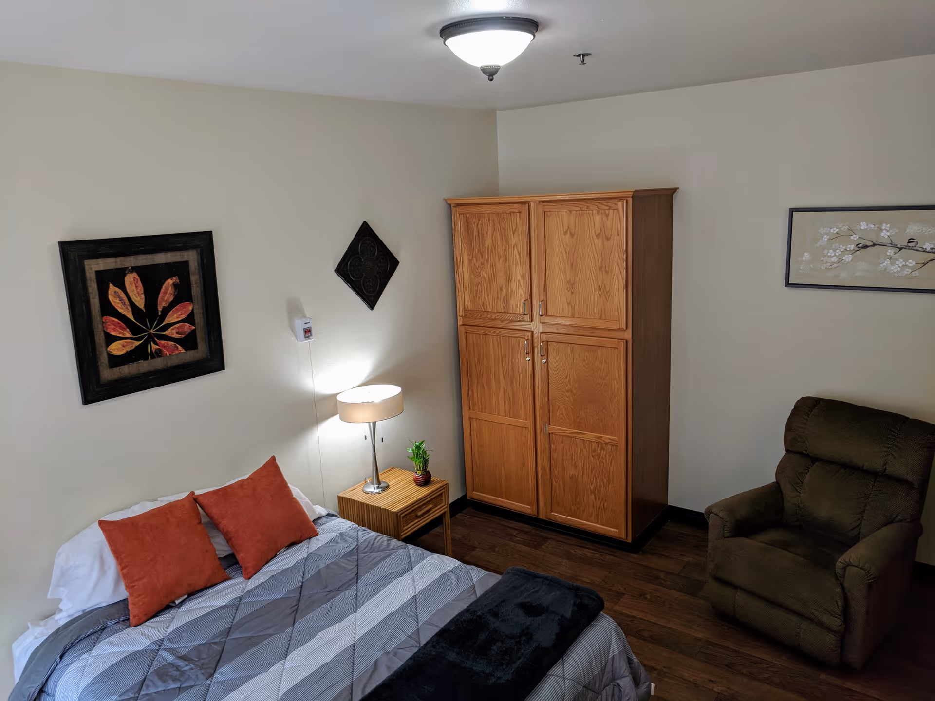 A cozy bedroom with a bed featuring a gray and white striped comforter and two red pillows. Next to the bed is a wooden nightstand with a lamp and a small plant. There is a large wooden wardrobe against the wall and a dark brown recliner chair. The walls are decorated with three pieces of artwork, and the floor is wooden.