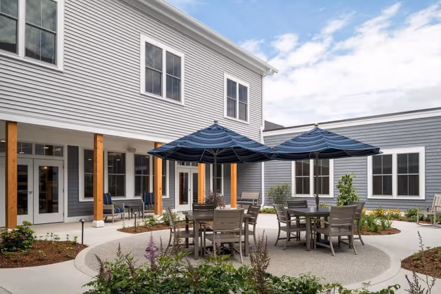 Outdoor courtyard area at Maple Ridge Lodge Assisted Living featuring round tables with chairs and large blue striped umbrellas. The courtyard is surrounded by a two-story building with gray siding and white trim, wooden pillars, and landscaped garden beds.