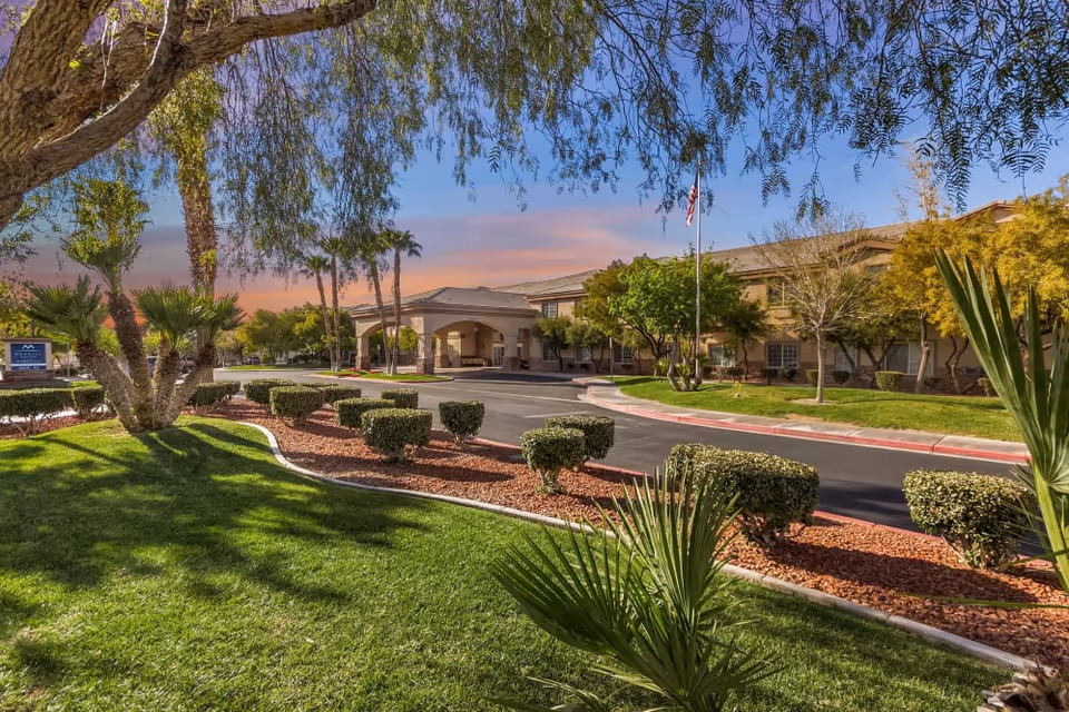 Exterior view of Truewood By Merrill, Henderson senior living facility at sunset with a driveway, manicured bushes, green lawn, palm trees, and an American flag in front of the building.