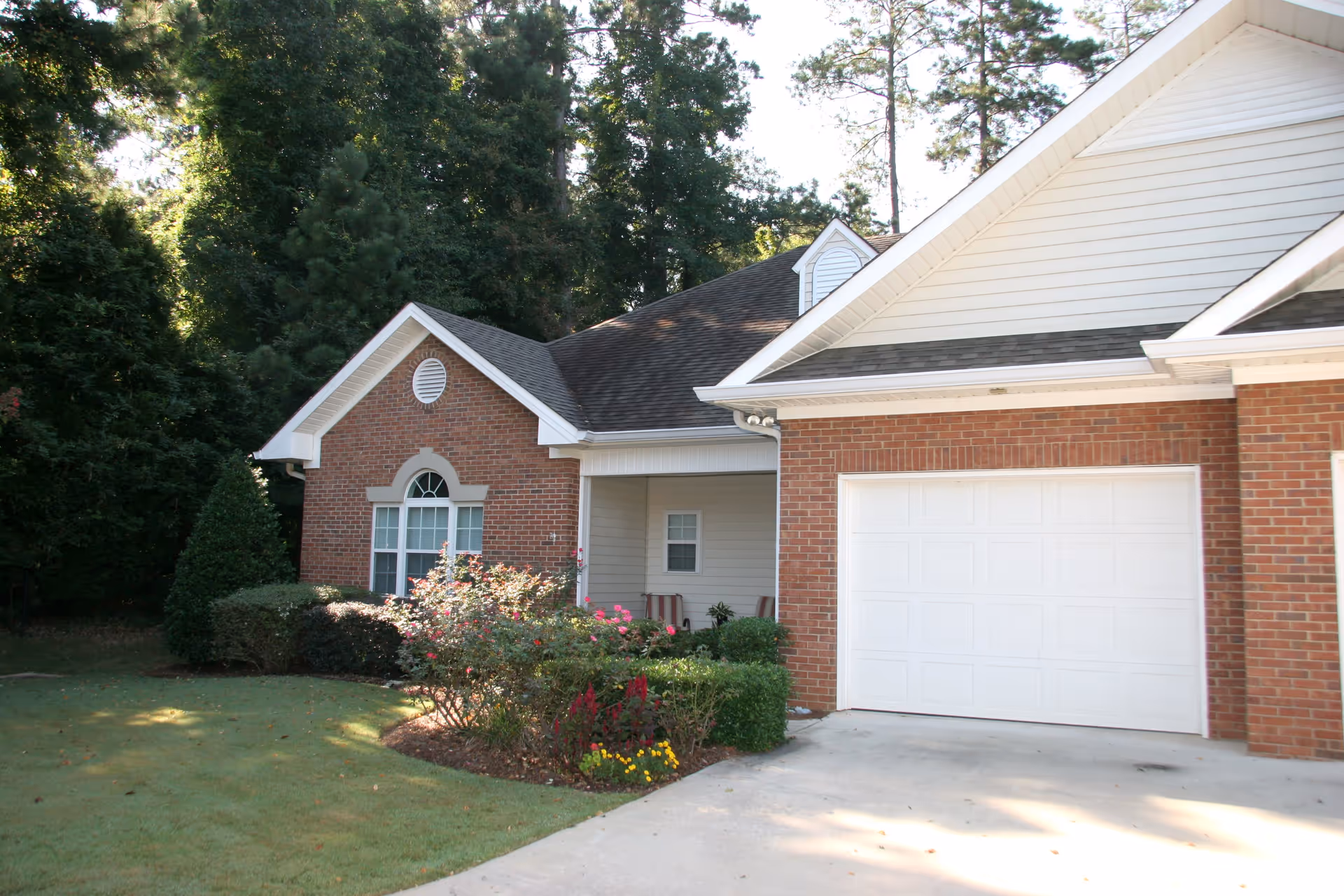 Exterior view of a single-story brick house with a white garage door, a small porch area, and a well-maintained garden with bushes and flowers in front. Tall trees are visible in the background.
