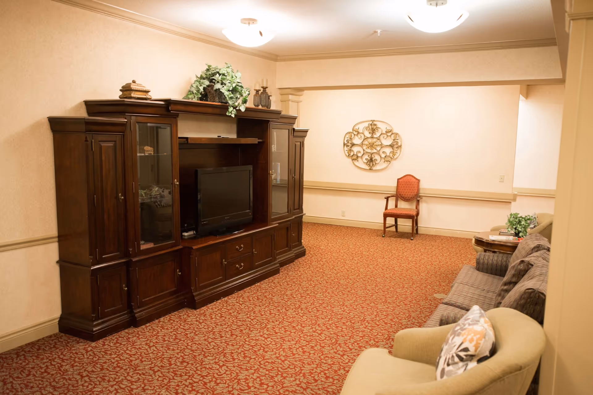 Carpeted lounge with a dark wood entertainment center and TV, upholstered chairs and sofas, and decorative wall art on the far wall.