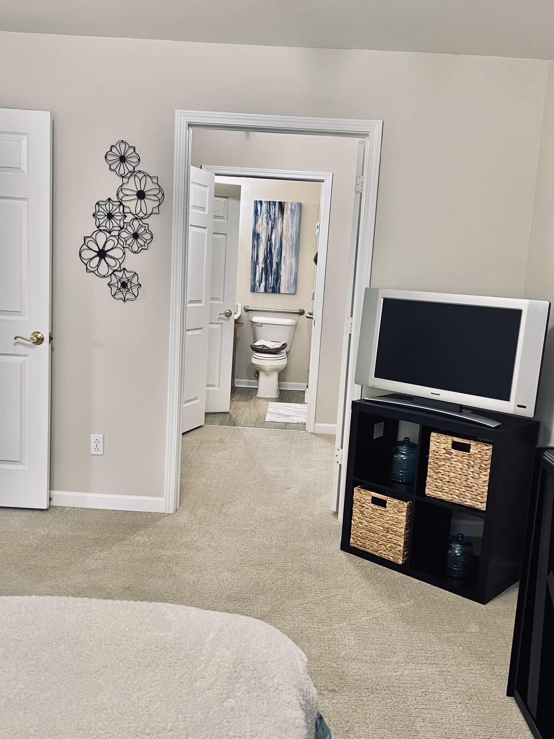 View of a bedroom with beige carpet and light-colored walls. A TV sits on a black cubed shelf with woven baskets and decorative jars. The door to a bathroom is open, showing a toilet, a towel on the toilet seat, a grab bar, and a blue and white abstract painting on the wall. A decorative metal wall art piece with flower designs is mounted on the wall near the door.