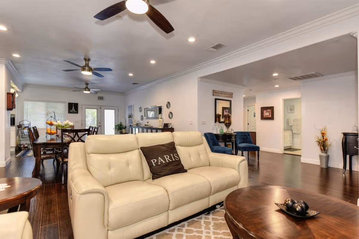 A spacious and well-lit living room area with a cream-colored leather sofa featuring a pillow with the word 'PARIS'. The room has dark wooden floors, ceiling fans with lights, and a wooden coffee table with decorative items. In the background, there is a dining area with a wooden dining table and chairs, and various decorative elements on the walls and surfaces.