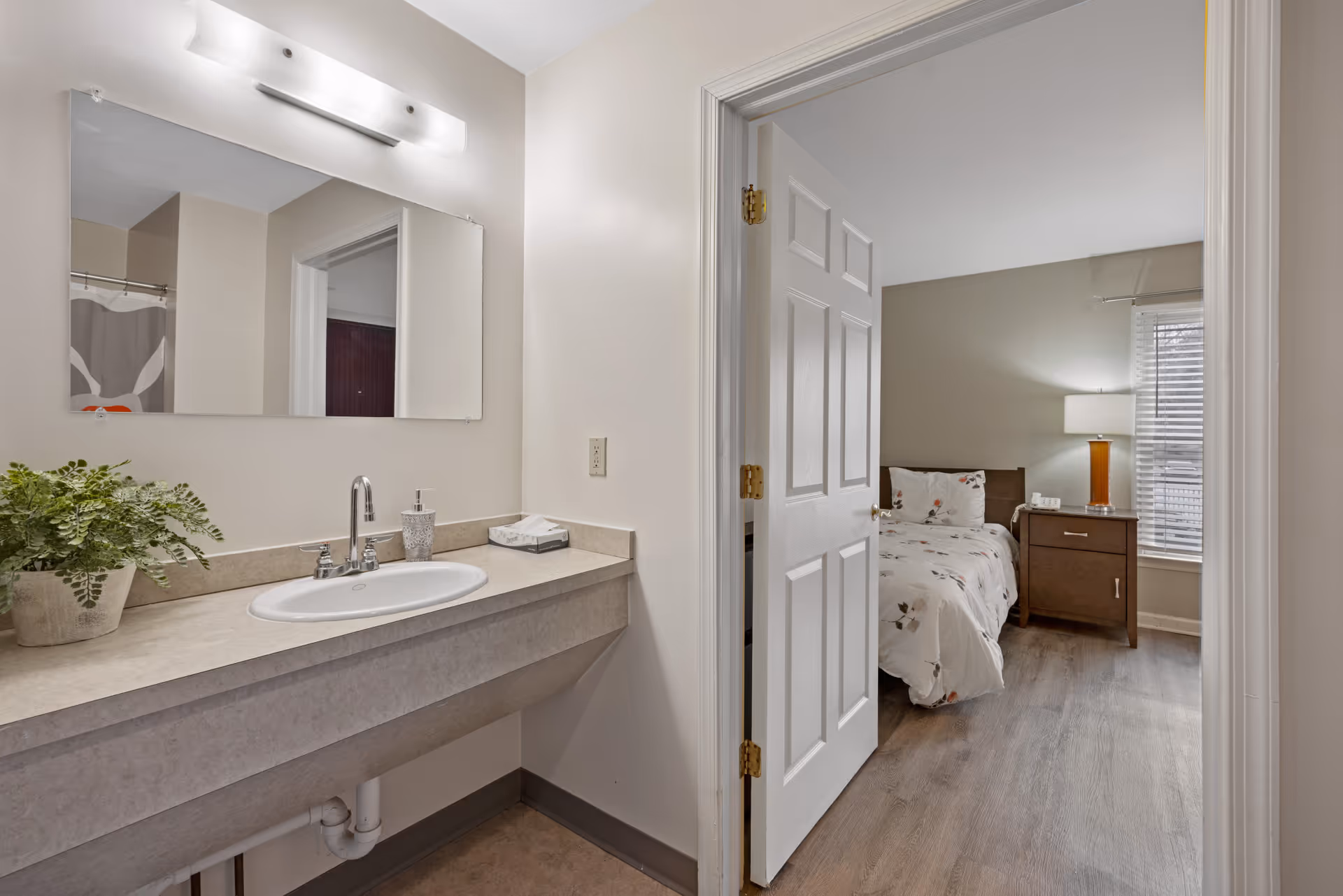 View of a bathroom sink area with a large mirror, soap dispenser, tissue box, and a potted plant on the counter. Through an open door, a bedroom is visible with a bed, nightstand, lamp, and window with blinds.