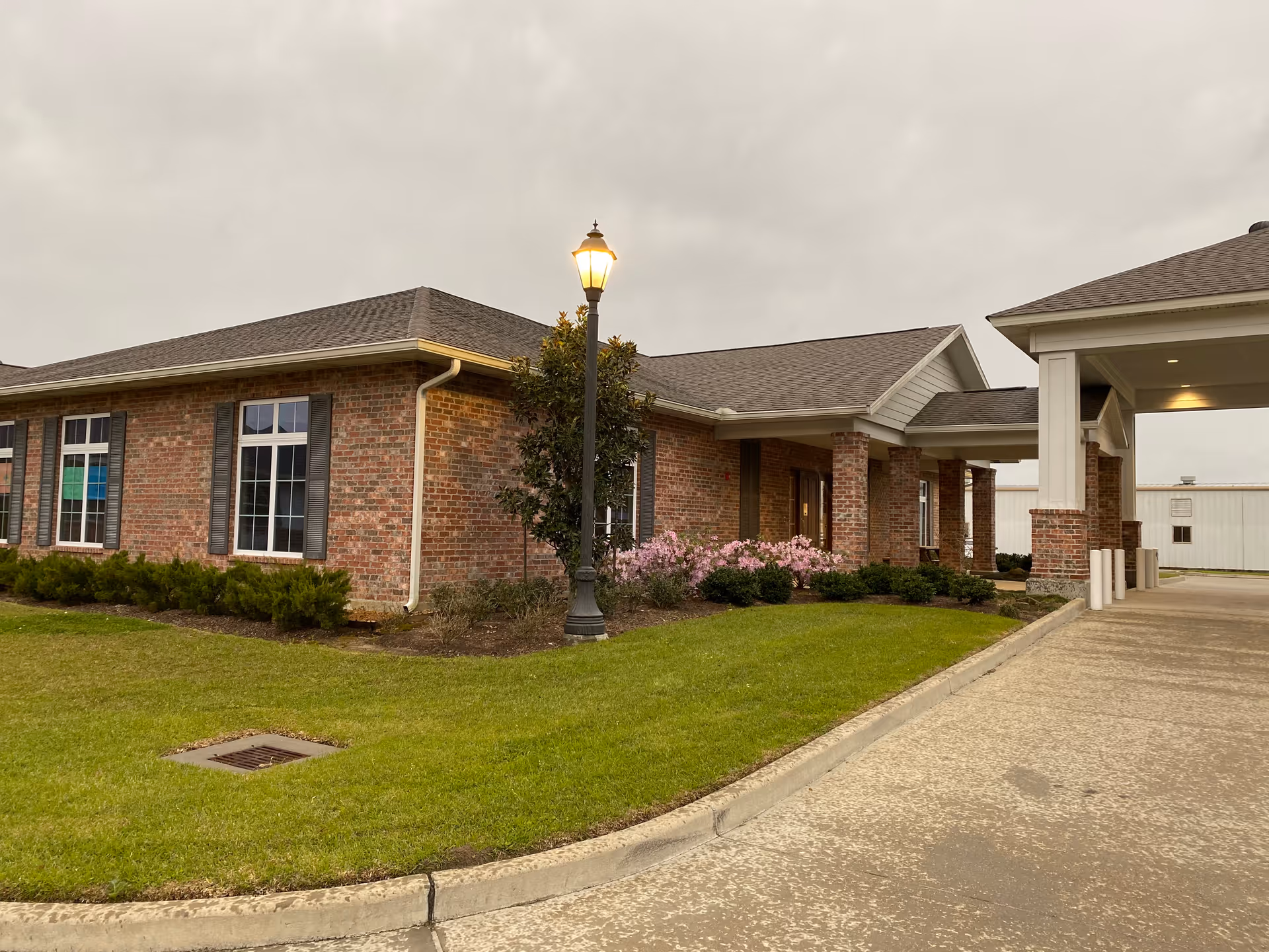 Exterior view of a brick building with a covered entrance, a lit streetlamp, green grass, and some bushes and flowers under a cloudy sky.