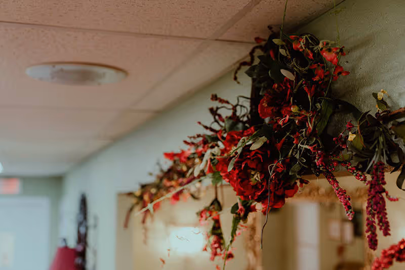 Close-up view of a decorative floral arrangement with red flowers and green leaves hanging on a wall inside a building hallway.