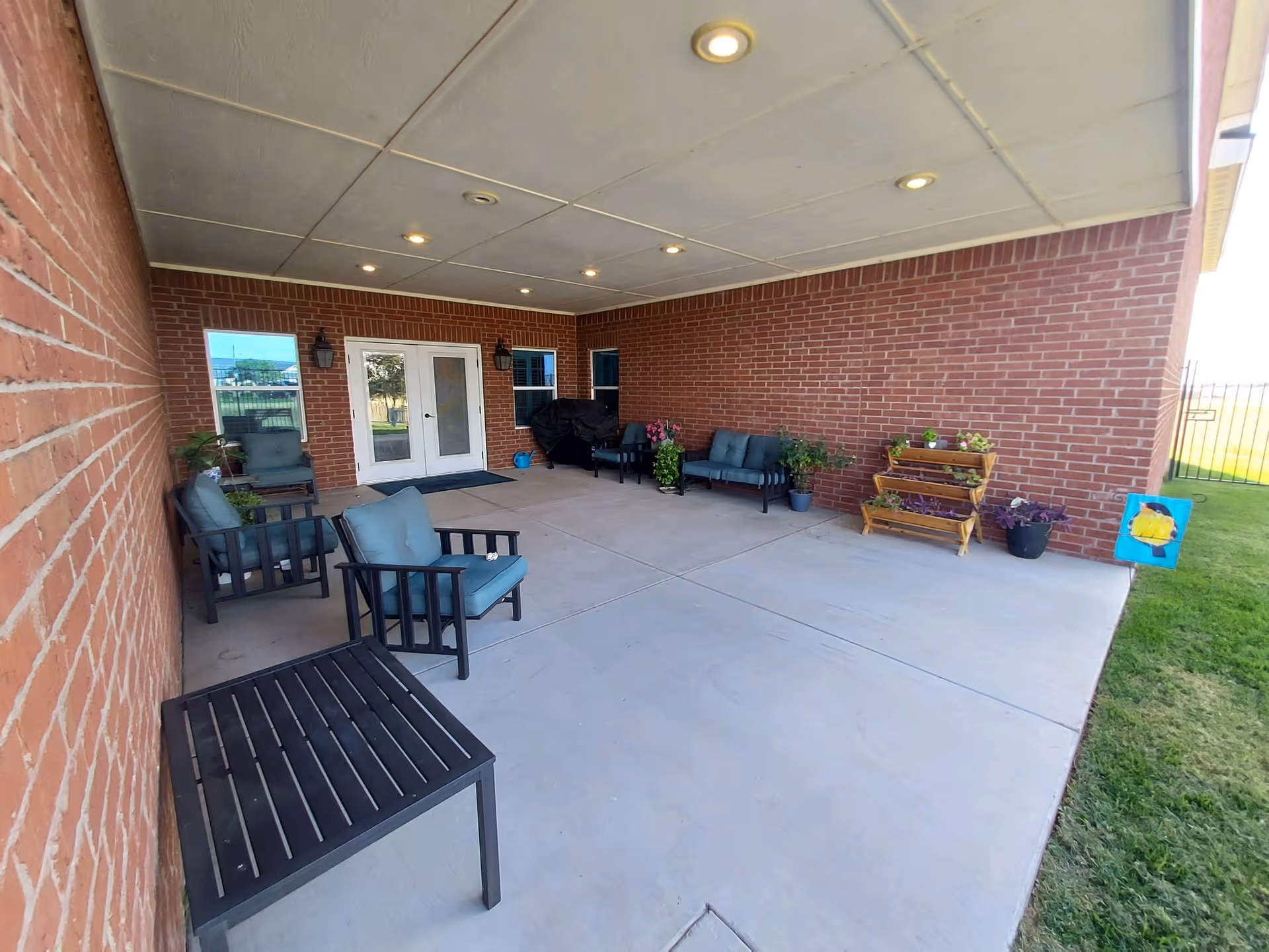 Covered outdoor patio area with brick walls, concrete floor, and ceiling lights. The patio is furnished with several cushioned chairs, a small table, potted plants, and a wooden plant stand. Double glass doors lead inside the building, and a grassy area is visible to the right.