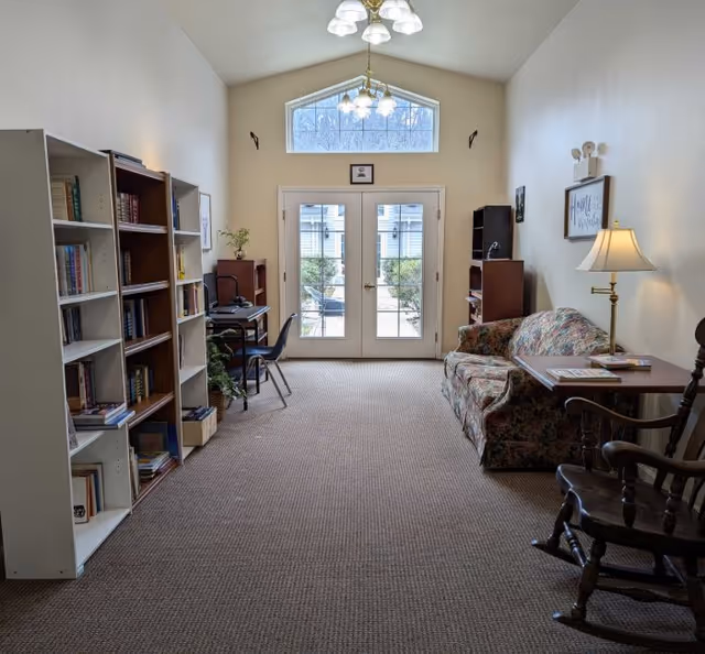 A cozy room with a floral-patterned sofa, a wooden rocking chair, a side table with a lamp, bookshelves filled with books, a desk with a chair, and double glass doors leading outside. The room has a high ceiling with a window above the doors and two ceiling light fixtures.