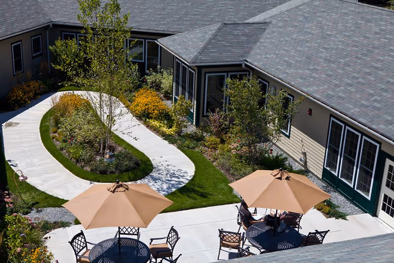 Landscaped courtyard with a winding concrete path, garden beds, and patio tables with umbrellas beside a single-story building.