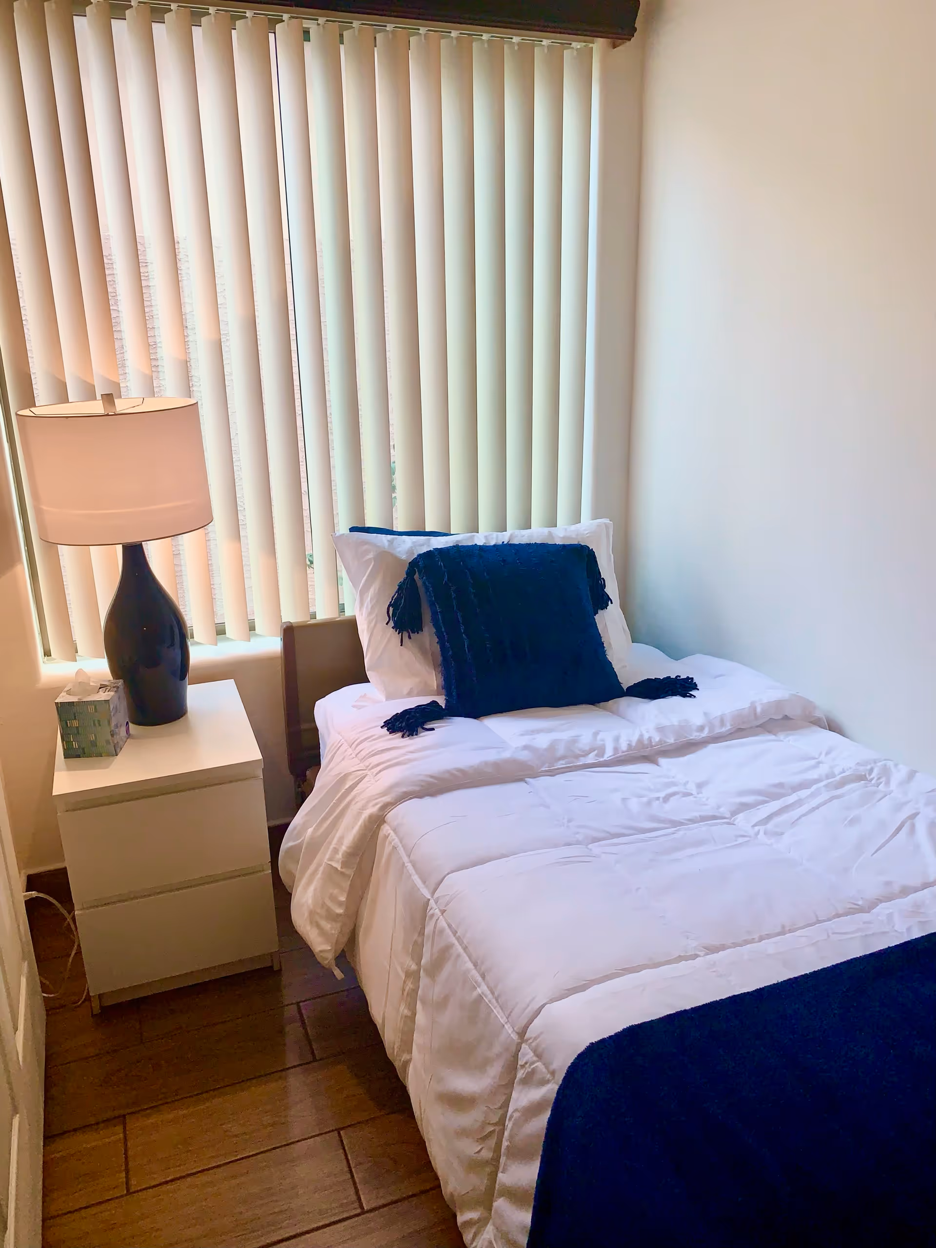 A small bedroom with a single bed made with white bedding and a dark blue throw blanket and pillow. Next to the bed is a white nightstand with a black lamp and a box of tissues. Vertical blinds cover the window behind the bed.