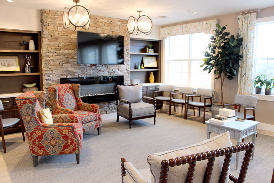 A bright and cozy living room with patterned armchairs, a stone fireplace with a mounted flat-screen TV above it, wooden shelves with decorative items, a row of chairs along the window, a small table with books, and large windows with floral curtains letting in natural light.