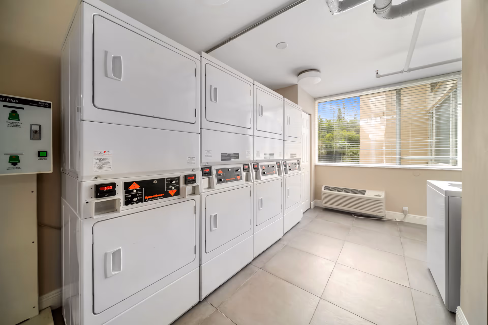 Laundry room with multiple stacked white washers and dryers along one wall, a large window with blinds letting in natural light, and a beige wall with a heating/cooling unit beneath the window.