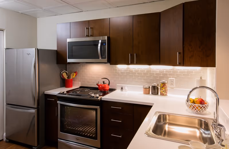 Modern kitchen with stainless steel refrigerator, oven, and microwave. Dark wood cabinets with silver handles, white countertops, and a white subway tile backsplash. A red kettle on the stove, a red utensil holder with cooking utensils, jars with ingredients, a paper towel holder, and a fruit basket with apples and bananas on the counter. A stainless steel sink with a curved faucet is also visible.