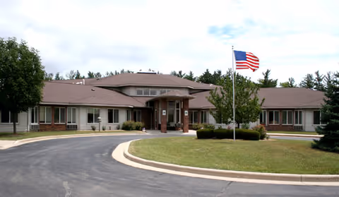Front exterior view of a single-story senior living facility building with a curved driveway, an American flag on a flagpole, and surrounding greenery including trees and bushes under a cloudy sky.