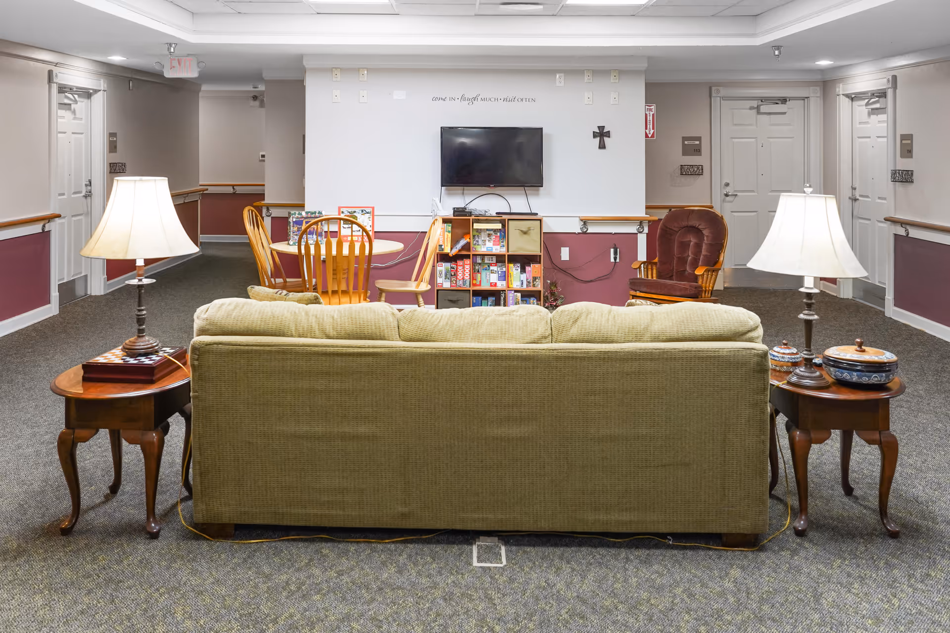 A communal lounge in a senior facility with a green couch facing a wall-mounted TV, side tables with lamps, a small dining table, and bookshelves.