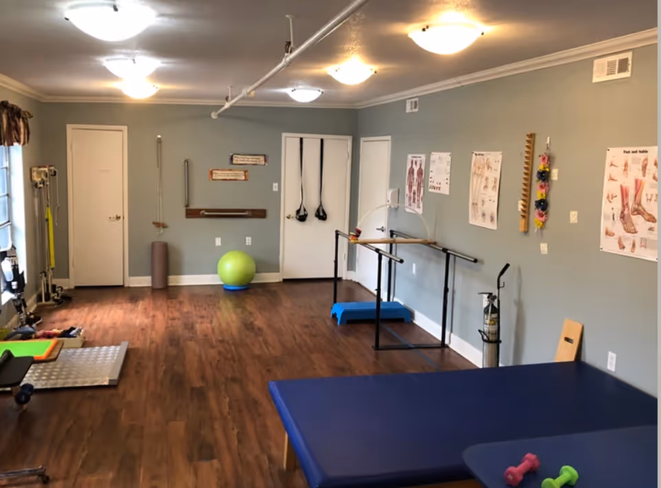 A physical therapy or rehabilitation room with wooden flooring, exercise equipment including a green exercise ball, resistance bands hanging on the wall, parallel bars for walking practice, anatomical charts on the walls, and a blue therapy table with small dumbbells on it.