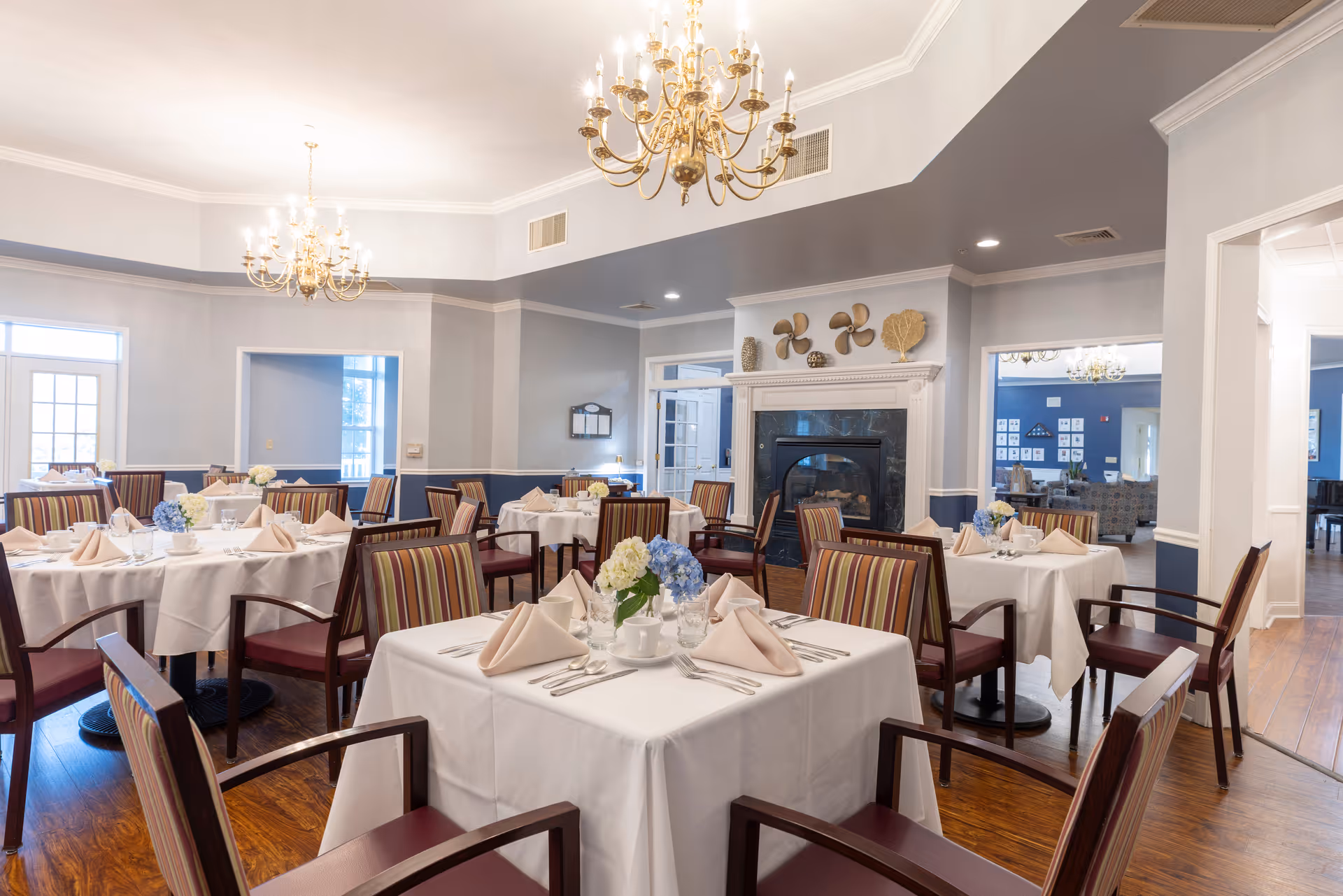 A bright and elegant dining room with multiple tables covered in white tablecloths, each set with beige napkins, silverware, cups, and floral centerpieces. The room features wooden floors, striped cushioned chairs, two gold chandeliers, and a fireplace decorated with nautical-themed ornaments.