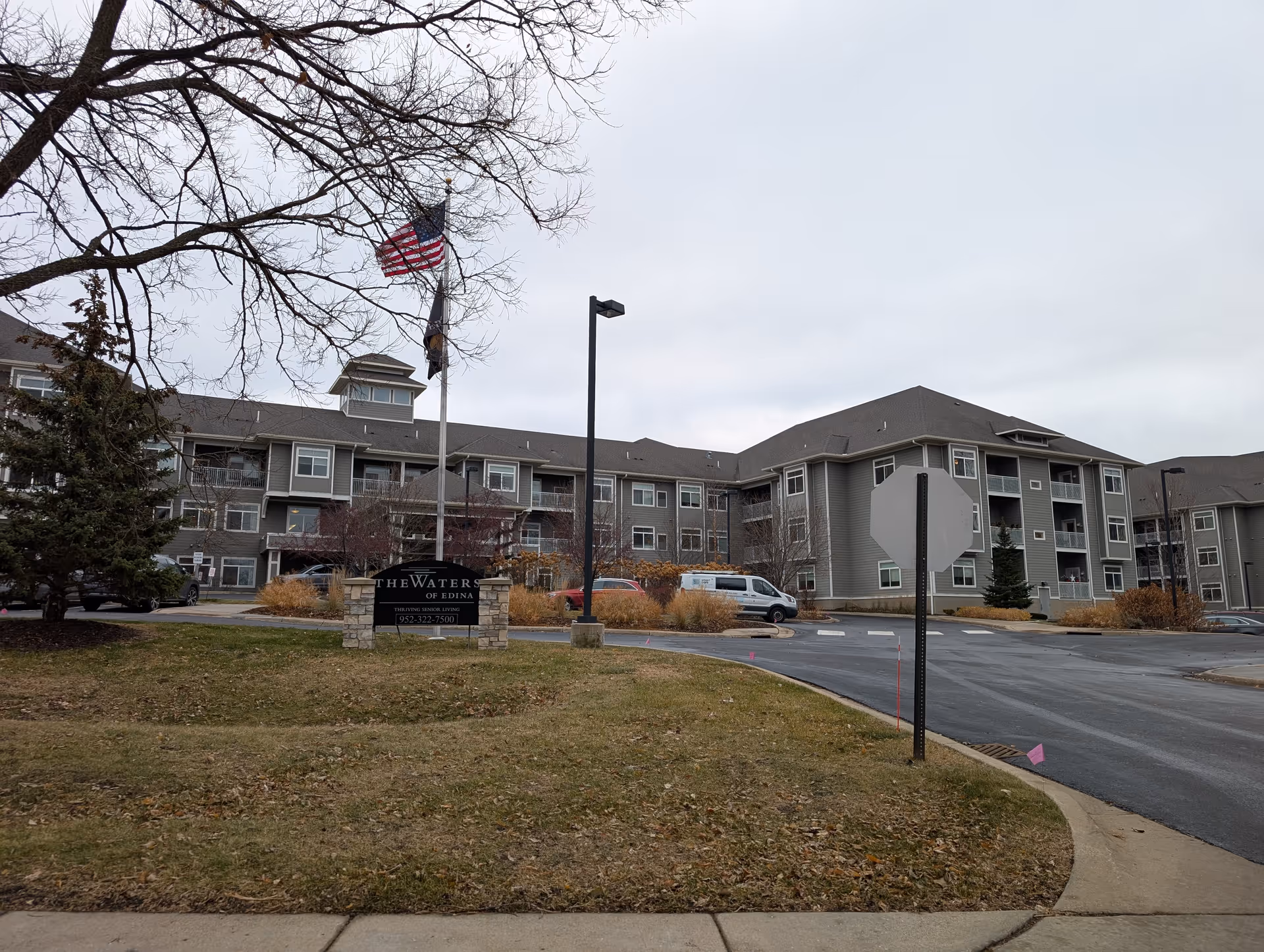 Exterior view of The Waters on 50th senior living facility showing a large multi-story building with gray siding, several windows, and balconies. There is a sign in front of the building with the facility name and phone number. An American flag and another flag are flying on flagpoles near the sign. The scene includes a paved driveway, some parked vehicles, leafless trees, and a cloudy sky.