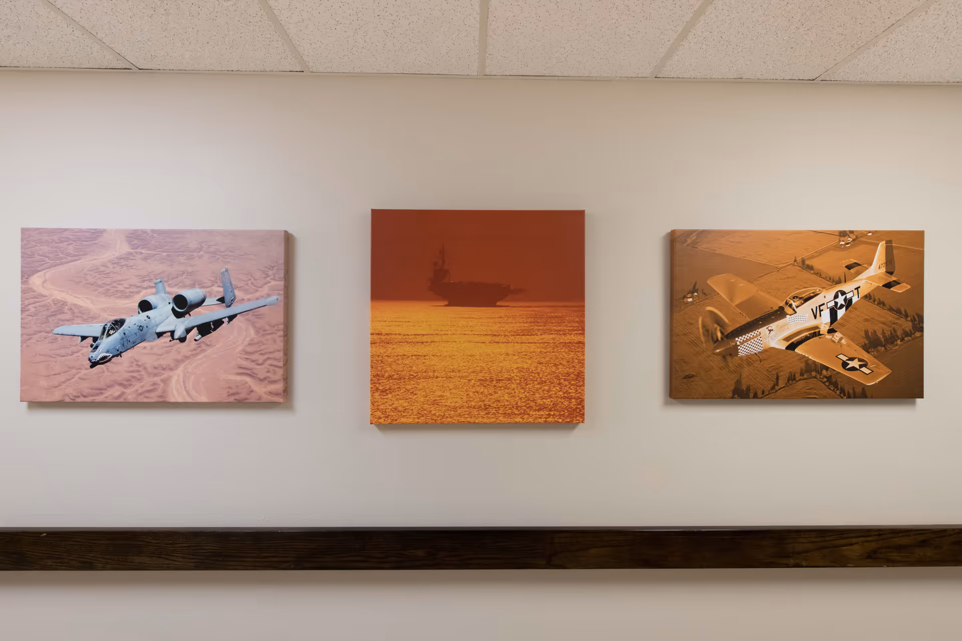 Three framed pictures on a beige wall above a wooden handrail. The left picture shows a military aircraft flying over a desert landscape. The middle picture depicts an aircraft carrier silhouetted against an orange sunset over the ocean. The right picture features a vintage military airplane flying over farmland.