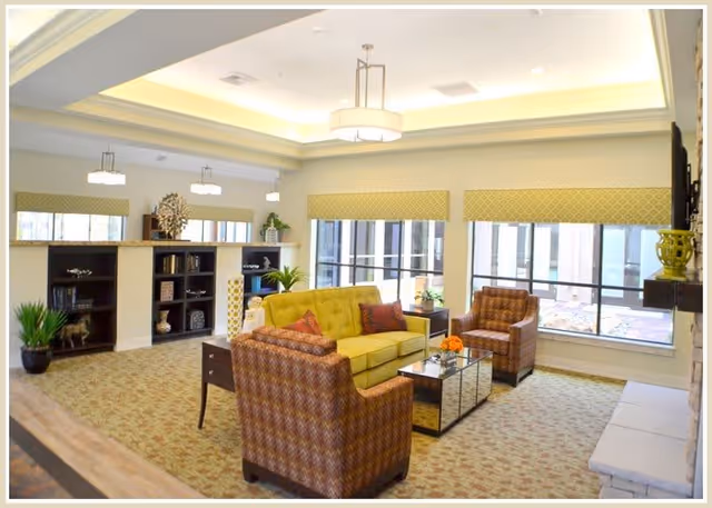 Bright communal lounge with upholstered chairs and a sofa arranged around a glass coffee table, bookshelves, and large windows.
